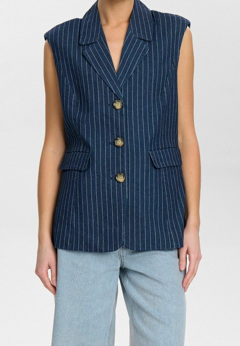 Blazer sans manches bleu marine à fines rayures avec trois boutons et poches à rabat à l'avant, porté sur un jean bleu clair.