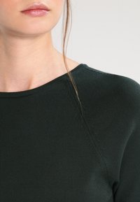 Pull en maille vert foncé avec un col arrondi, des détails de couture subtils et une coupe ajustée qui met en valeur la zone des épaules.