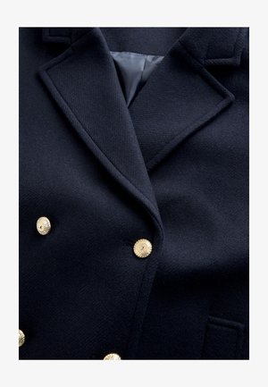 Tengerészkék gyapjú blézer aranyszínű gombokkal, bevágott gallérral és két elülső zsebfedővel. Texturált szövet sima belső béléssel.