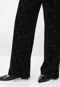 Schwarze Hose mit einem strukturierten Damastmuster; weiter Beinansatz, glattes Material und kombiniert mit spitzen schwarzen Ballerinas.