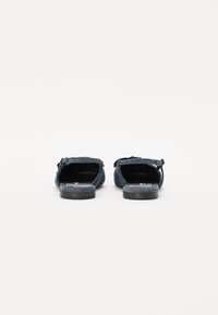 Chaussures en denim à enfiler avec une semelle en caoutchouc noir plat, dos ouvert et accents de boucle. Elles présentent un dessus texturé bleu et une semelle intérieure plate.