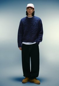 Marinblå texturerad stickad tröja, vit t-shirt under, lösa svarta byxor och gula skor. Keps med röda accenter på en modell mot en gradient bakgrund.