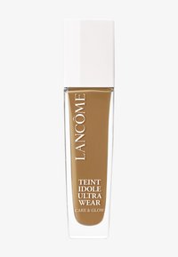 Fond de teint liquide marron dans une bouteille en verre transparent avec un capuchon blanc. L'étiquette indique "LANCÔME TEINT IDOLE ULTRA WEAR CARE & GLOW."