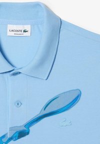 Vaaleansininen polo-paita, jossa on teksturoitu kangas, jossa on pieni Lacoste-logo ja kontrastinvärinen sininen lusikan graafinen kuvasto.