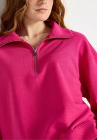Kvinde med rødt hår iført en lys pink sweatshirt med kvart lynlås, der viser lynlåsdetaljer og tekstureret stof på en lys baggrund.