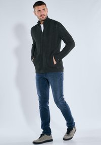 Dunkelgrüner gestrickter Zip-up-Jacke mit Fischgrätenmuster, kombiniert mit blauen Jeans und grauen Sneakers. Die Jacke hat seitliche Taschen.