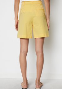 Vrouw in hoge gele shorts met achterzakken, staand op blote voeten tegen een eenvoudige witte muur.