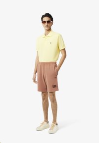 Camisa polo amarilla clara con logo, pantalones cortos rosas con un parche y zapatillas blancas. Modelo con gafas de sol marrones de pie contra un fondo uniforme.