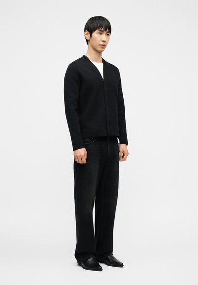 Άνδρας στέκεται φορώντας μαύρη ζακέτα με φερμουάρ, λευκό T-shirt, μαύρα φαρδιά τζιν και μαύρα μυτερά παπούτσια, πάνω σε απλό λευκό φόντο.