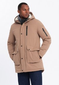 Parka beige con cappuccio foderato in pelliccia, bottoni frontali, tasche con cerniera nere e due grandi tasche anteriori con patta. Indossato sopra una sciarpa grigia.