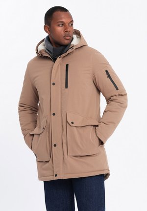 Beige parkajakke med pelsforet hætte, frontknapper, sorte lynlåslommer og to store frontlommer med klap. Båret over et gråt tørklæde.