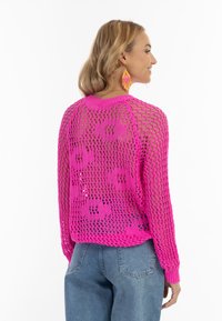 Rosa, gestrickter, verkürzter Pullover mit einem lockeren Netzdesign und floralem Muster. Verfügt über lange Ärmel und gerippte Bündchen. Kombiniert mit blauen Jeans.