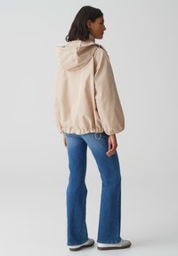 Giacca impermeabile beige con cappuccio, orlo elastico e maniche larghe. Abbinata a jeans a zampa d'elefante blu e sneakers grigie. Design semplice e funzionale.