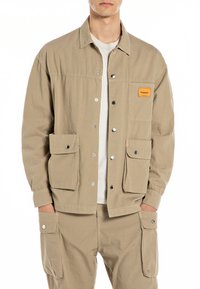 Beige lightweight jacket with a collared design, snap buttons, and two front pockets. Features a small orange label on the left chest.