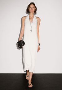 Weißes Halfterkleid mit fließender Silhouette. Kombiniert mit einer strukturierten schwarzen Clutch und silberner Schmuck, getragen mit Flip-Flops.