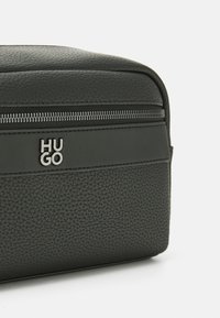 Svart teksturert lærpung med horisontal glidelås og sølv "HUGO"-logo på en glatt lærstripe.