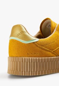Sneaker gialla in suede con una suola texturizzata e ondulata; presenta dettagli in pelle dorata e un bordo verde menta vicino al tallone per creare contrasto.