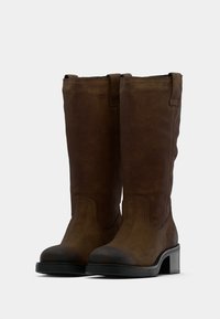 Bottes hautes en daim marron avec une finition texturée, bout rond, talon carré et languettes en haut pour un enfilage facile.