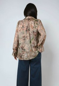 Frau mit langen dunklen Haaren, die eine durchsichtige Bluse mit floralen Mustern in gedämpften Tönen und eine dunkle, weit geschnittene Jeans trägt, steht mit einer Hand auf der Hüfte.