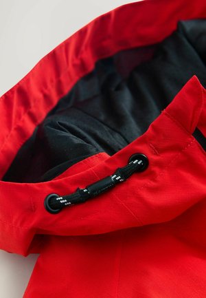 Rød jakke med en tekstureret yderside. Indersiden har en sort foring, en snor med justeringsstop, og diskrete perforeringer for ventilation.