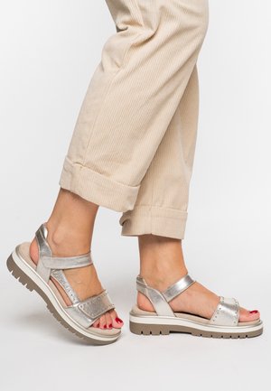 Silver sandals with a chunky sole, featuring a mix of smooth and textured straps, and decorative studs along the upper. Worn with rolled beige pants.
