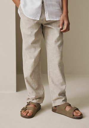 Person, die beigen Leinenhosen, taupefarbene, mit Schnallen versehene Sandalen und ein weißes Hemd trägt, vor neutralem Hintergrund steht.