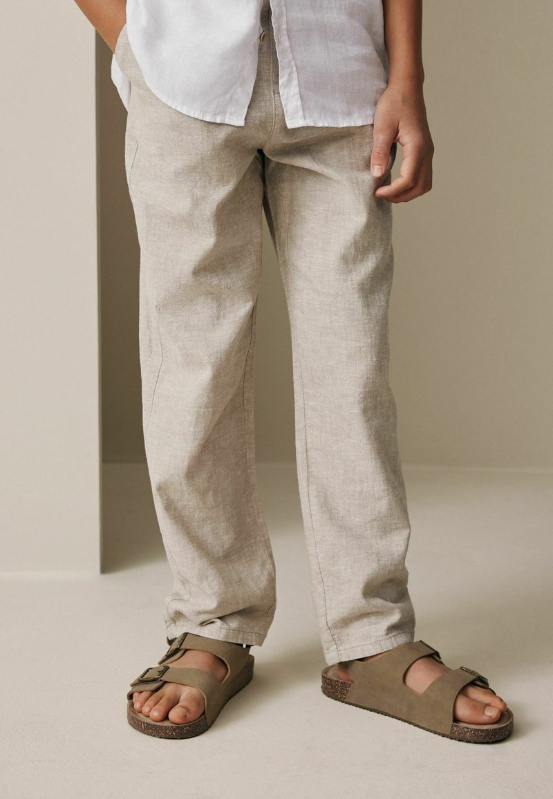 Persona de pie con pantalones de lino beige, sandalias taupe con hebilla y camisa blanca sobre un fondo neutro.