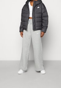 Chaqueta de plumas negra con capucha, pantalones de chándal grises y zapatillas blancas. La chaqueta presenta el logo de Nike; los pantalones de chándal son de pierna ancha con cordón.