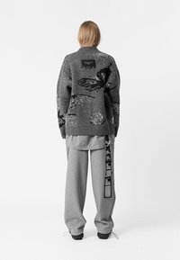 Suéter gris de punto con patrones abstractos, acentos negros y una etiqueta en la parte trasera; combinado con pantalones deportivos gris claro que tienen una etiqueta lateral.