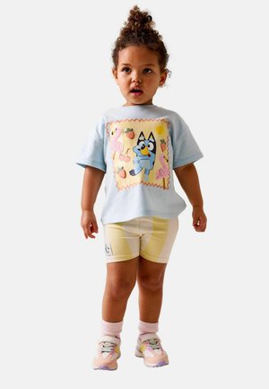 Tricou albastru deschis cu un desen animat colorat, asortat cu pantaloni scurți cu dungi galbene și adidași în nuanțe pastelate.