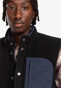 Black fleece vest with a navy pocket, snap button closure, and high collar, layered over a checkered shirt in neutral tones.