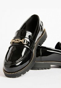 Mustad nahast mustad loafers, millel on kuldne ahel ja studidega tald. Disain on lame, ümara nina ja tekstuuriga detailidega.