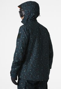 Kurtka śniegowa w ciemnoniebieskiej podstawie z wielokolorowym, abstrakcyjnym wzorem, regulowanym kapturem i teksturowanym materiałem z przymocowaną naszywką.