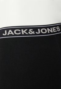 Černý bavlněný pásek s logem "JACK & JONES" v šedé barvě, obsahuje kontrastní bílou pruh podél horního okraje, detailní pohled na texturu látky.
