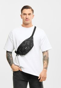 Marsupio mimetico nero con più scomparti con zip, tracolla regolabile, texture liscia e logo discreto sul davanti.