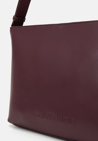 Burgundy leather handbag with embossed "Calvin Klein" logo, featuring a smooth texture and sleek rectangular design.