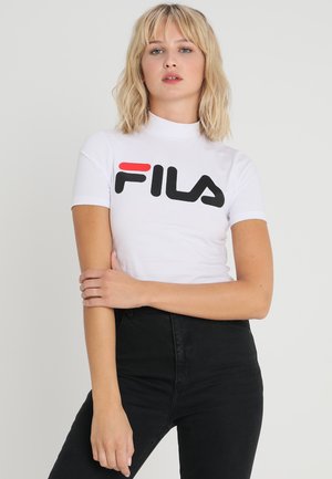 T-shirt blanc à col montant et à manches courtes, avec un logo FILA noir et rouge proéminent. Associé à un jean noir taille haute.