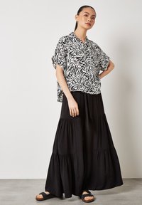 Schwarz-weiß gemusterte Bluse mit kurzen Ärmeln kombiniert mit einem langen schwarzen Stufenrock. Flache Sandalen runden das Outfit ab. Glatte Stoffstruktur.