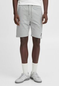 Shorts en coton gris avec une taille élastique et un cordon de serrage. Dotés de poches latérales et d'un petit logo sur la jambe gauche. Portés avec des chaussures de sport grises.