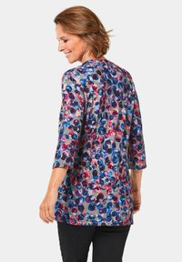 Multifarbene Tunika mit 3/4-Ärmeln, die ein gemustertes Design aus kreisförmigen Formen in Pink, Blau und Lila auf einem grauen Hintergrund zeigt.