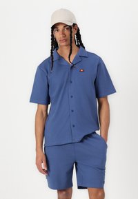 Chemise bleue à manches courtes avec col, ornée de boutons et d'un petit graphique sur la poitrine, associée à un short bleu assorti.