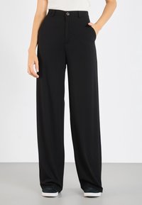 Femme portant un pantalon noir ample avec fermeture à bouton et fermeture éclair, poches latérales, associé à des baskets sombres et un haut blanc.