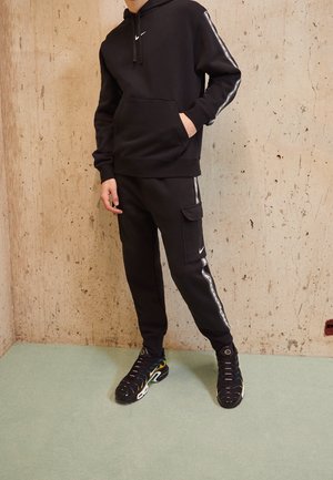 Osoba ubrana w czarną bluzę z kapturem Nike i spodnie cargo do biegania z białym paskiem i logo, stojąca przy strukturalnej beżowej ścianie i zielonej podłodze.