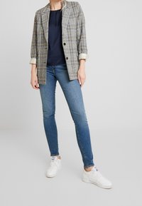 Kvinde iført en ternet blazer over en navy top, blå skinny jeans og hvide sneakers, stående imod en ensfarvet lys baggrund.
