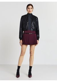 Giacca bomber in pelle nera, dolcevita grigia aderente, minigonna plissettata borgogna con cintura, calze nere e tacchi a punta borgogna.