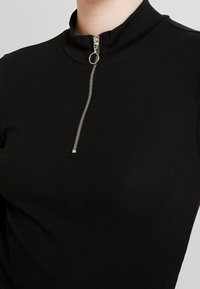 Svart ribbad topp med hög krage och kvartdragkedja fram, med detaljer i silver. Toppens design är strukturerad och figurnära.