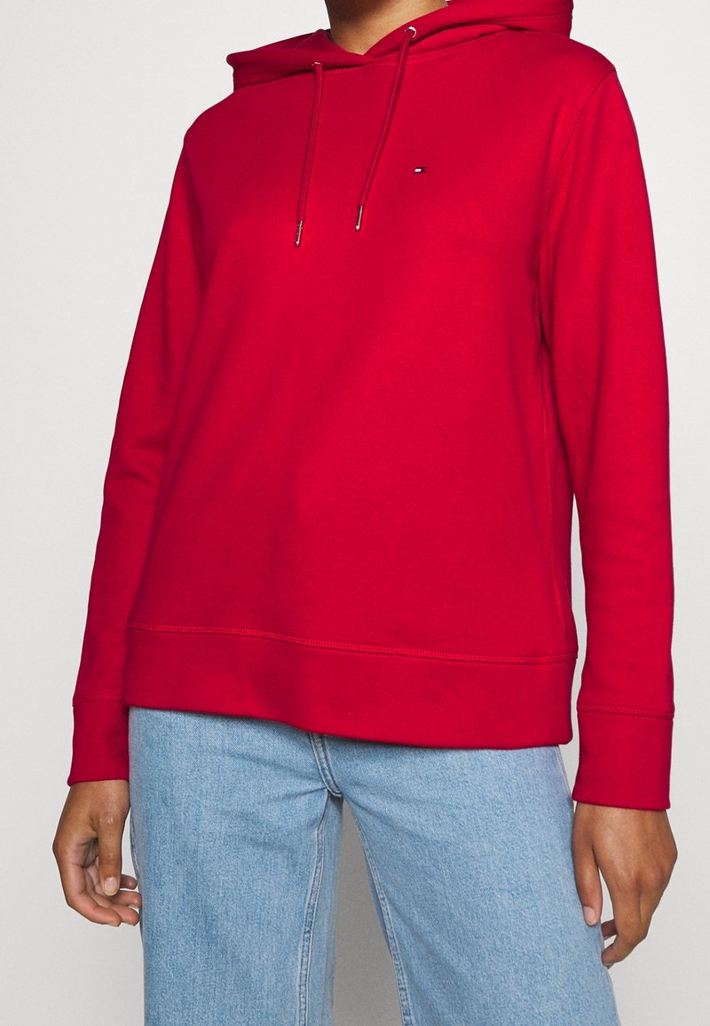 Personne portant un sweat à capuche rouge uni avec cordons à l'avant et un jean bleu clair, montrée du milieu du torse jusqu'aux mains.