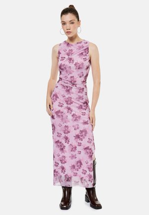 Vrouw met een mouwloze, roze bloemen maxi-jurk met zijsplit, gecombineerd met donkerbruine enkellaarzen, staand tegen een witte achtergrond.