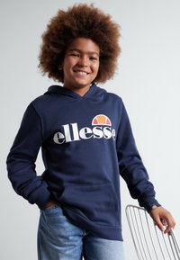 Marljivo plava dukserica s kangaroo džepom, koja se ističe odvažnim "ellesse" logotipom. Izrađena od teksturiranog materijala, sa rebrastim manžetama i kapuljačom.