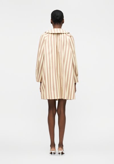 Γυναίκα με σκουρόχρωμη επιδερμίδα και μαλλιά σε κότσο φοράει μπεζ και ανοιχτό καφέ παλτό με κάθετες ρίγες και λευκά τακούνια, στέκεται με την πλάτη προς τα έξω σε λευκό φόντο.